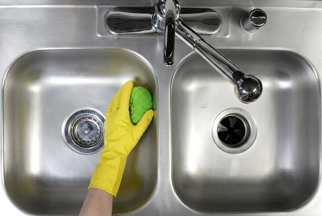 Person cleaning a stainless steel sink with a green sponge and yellow rubber glove.